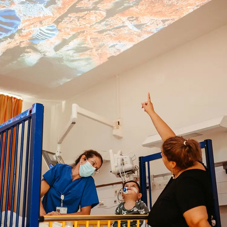 Een patiënt, zorgverlener en moeder kijken samen naar een rustgevende lichtprojectie van de Qwiek.up op de muur.
